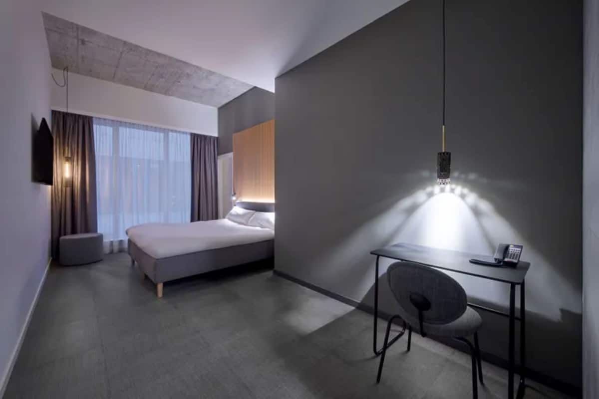 A modern hotel room with a bed, a desk with a chair, a mounted TV, and a large window with curtains.