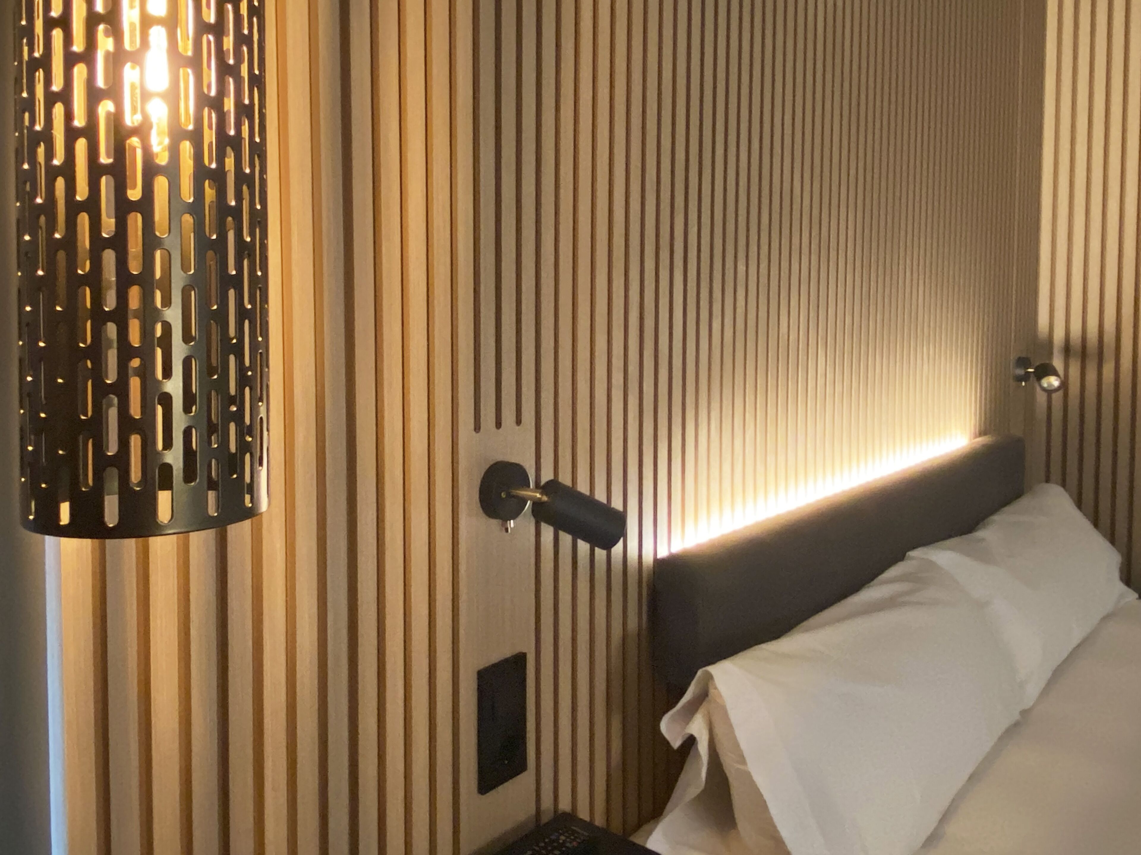 Una habitación de hotel con papel tapiz a rayas verticales, una cama con ropa de cama blanca, una mesita de noche negra y una lámpara de pared.