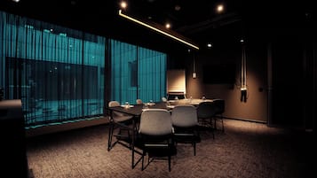 Une salle de conférence équipée d’une grande table, de chaises, d’un projecteur et d’une grande fenêtre donnant sur un immeuble.