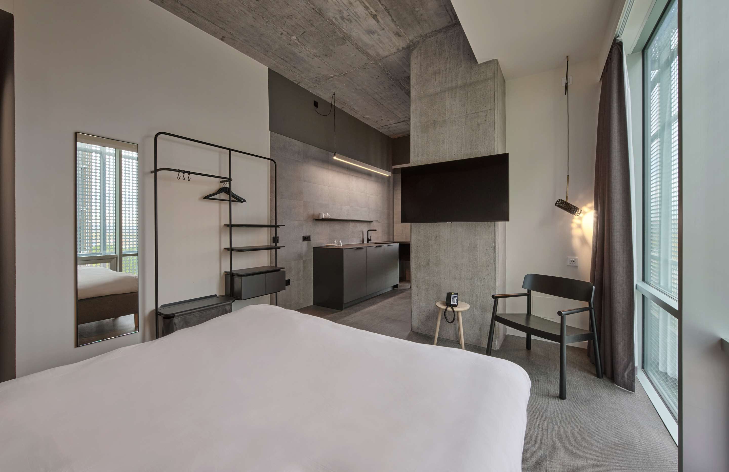 Een moderne hotelkamer met een groot bed, een klein bureau, een stoel, een aan de muur bevestigde televisie en een kitchenette op de achtergrond.
