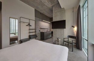 A modern hotel room with a large bed, a small desk, a chair, a TV mounted on the wall, and a kitchenette in the background.