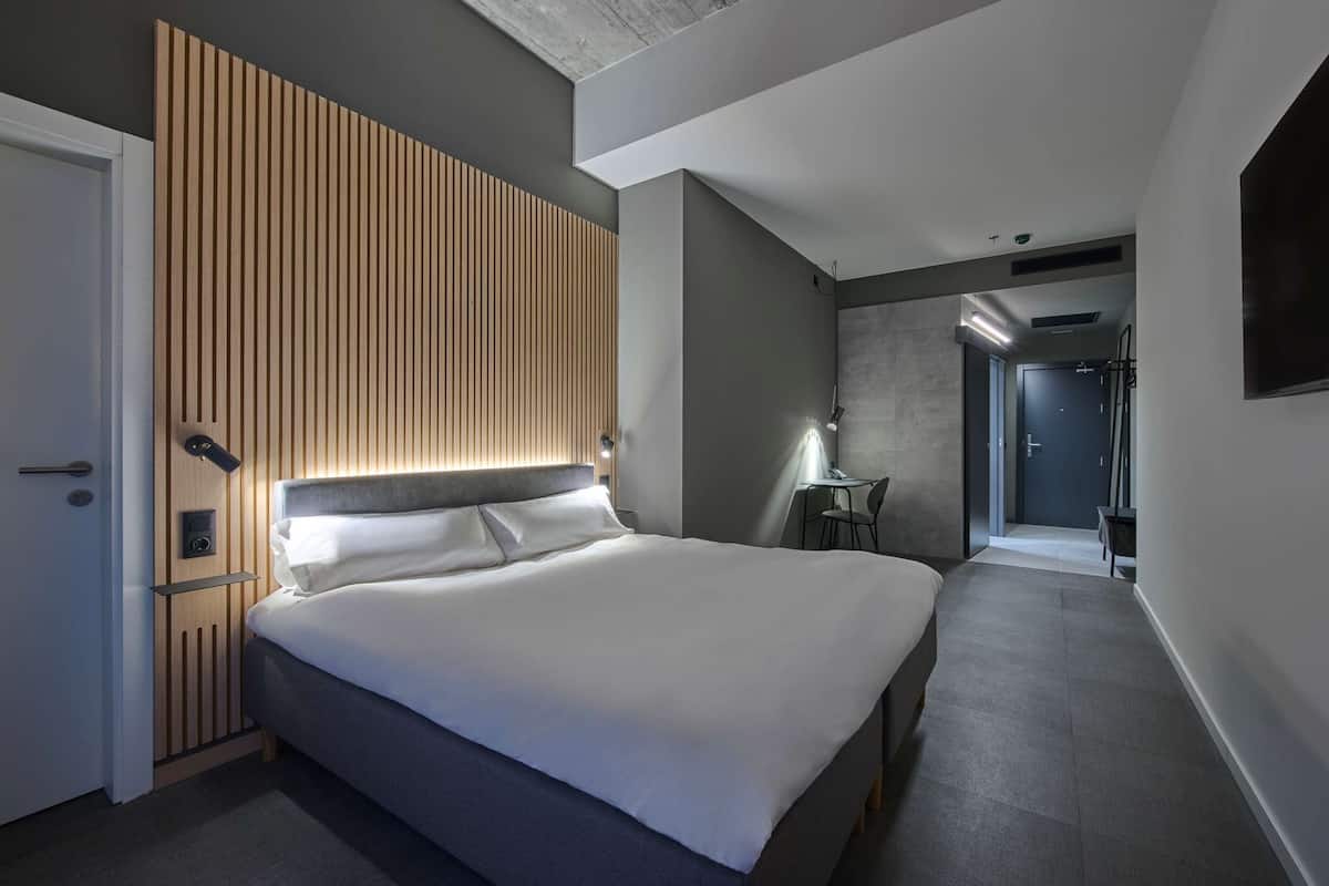 A modern hotel room with a large bed, a wooden slatted headboard, a desk, and a TV.