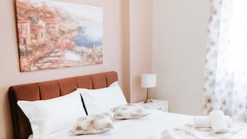 A neatly made bed with a floral bedspread, a white nightstand with a lamp, and a painting of a coastal scene on the wall.