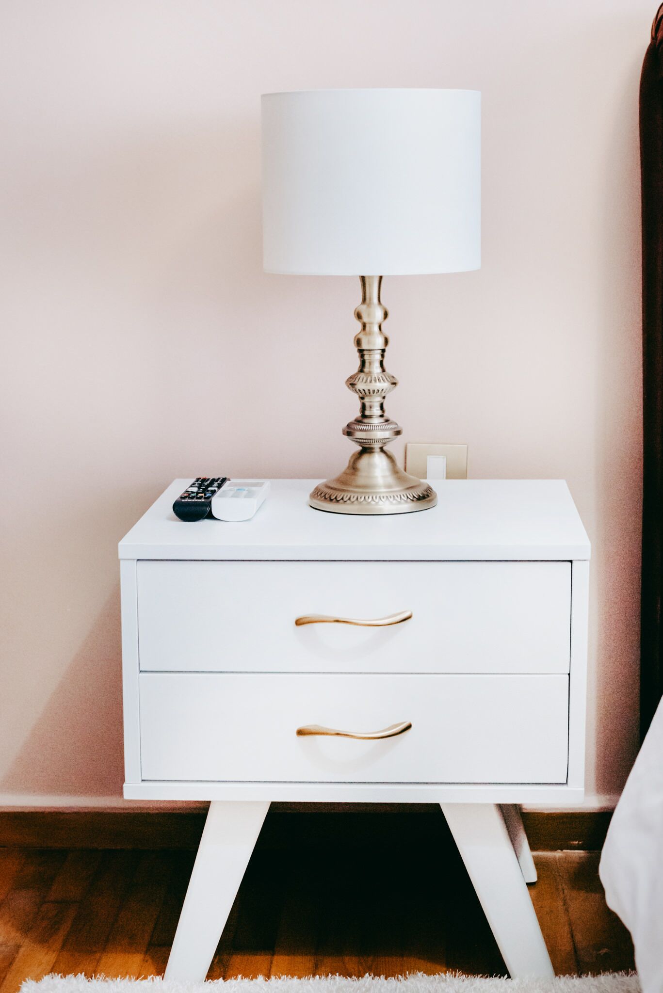 Een wit nachtkastje met een lamp, een afstandsbediening en een telefoon.
