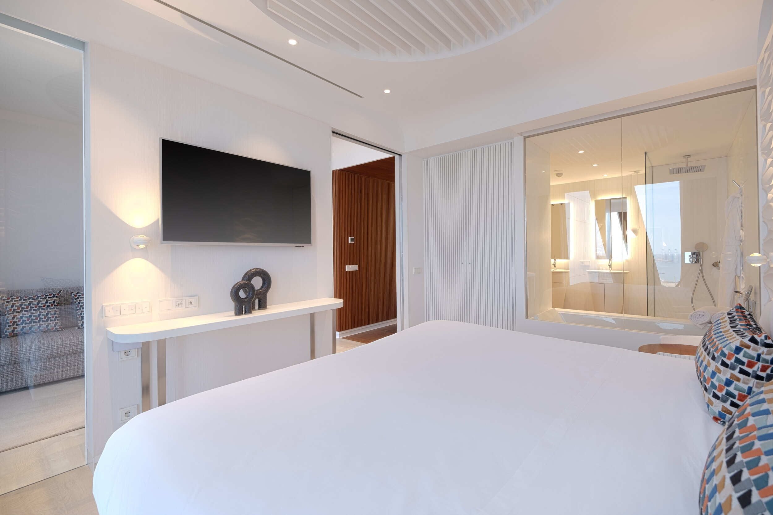 A modern bedroom with a large bed, a flat-screen TV mounted on the wall, a bedside table, and a glass shower area.