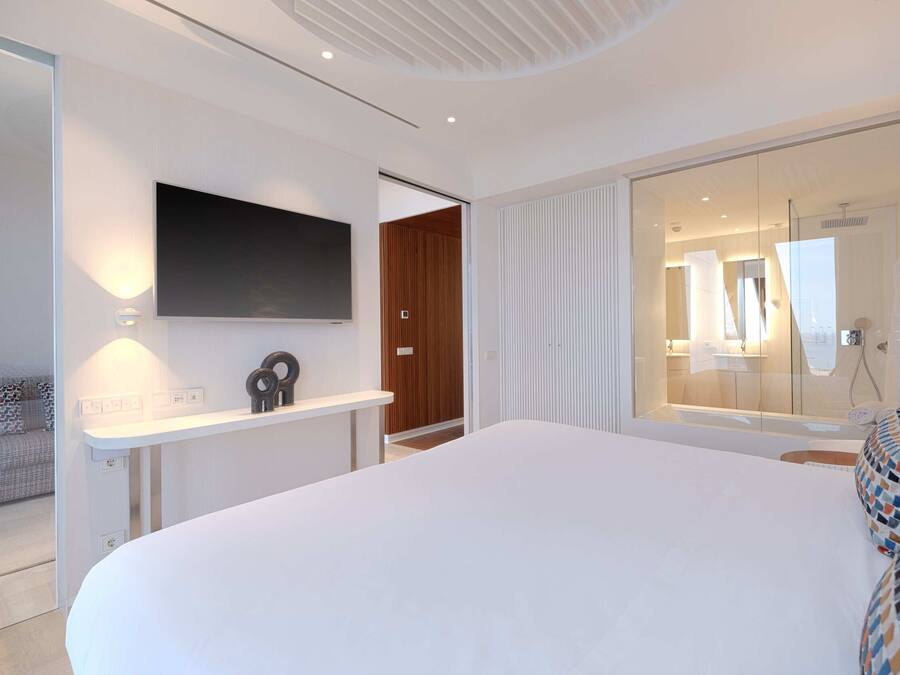A modern bedroom with a large bed, a flat-screen TV mounted on the wall, a bedside table, and a glass shower area.