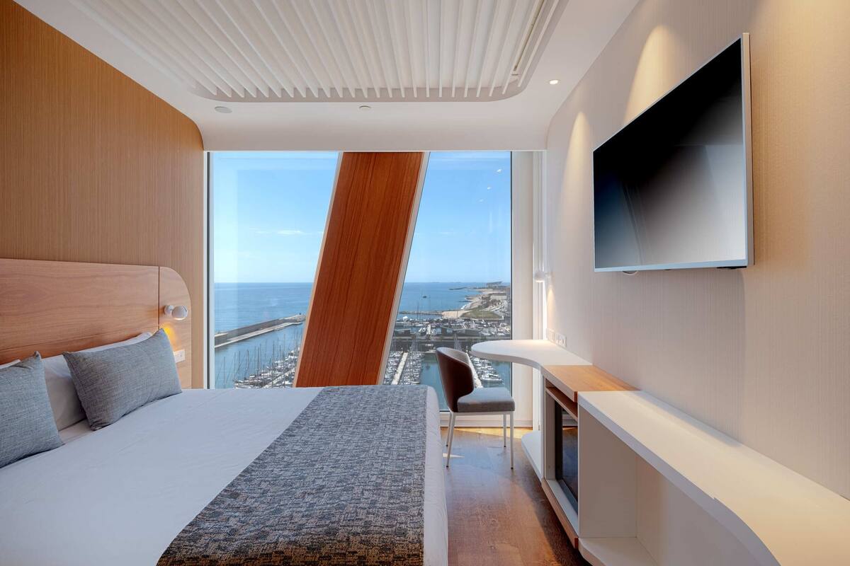 A modern hotel room with a large bed, a TV mounted on the wall, a desk, and a view of the city and sea.