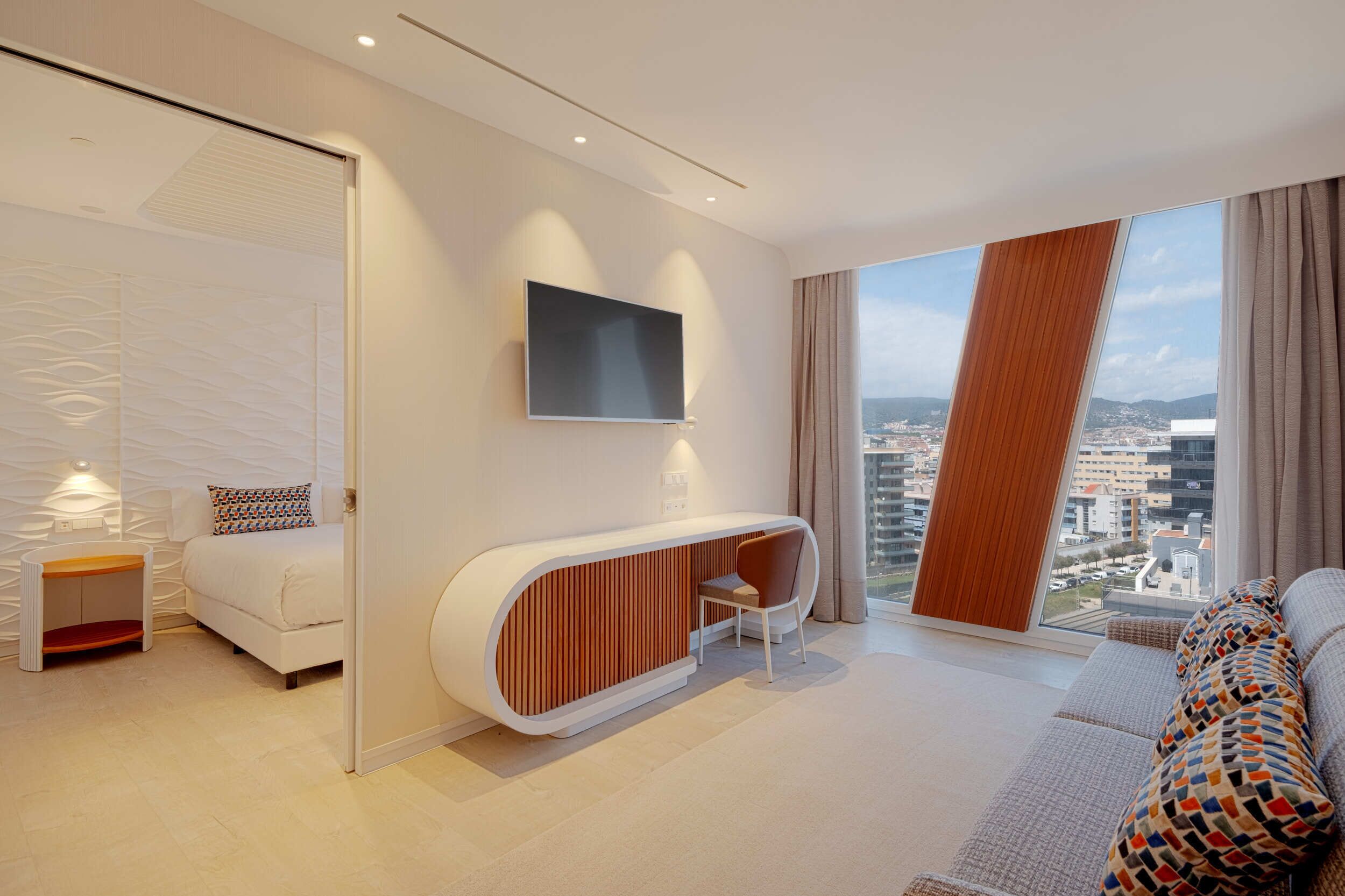 Un moderno cuarto de hotel con una cama, un televisor montado en la pared, una zona de estar con un panel de madera y un amplio ventanal con vistas a la ciudad.