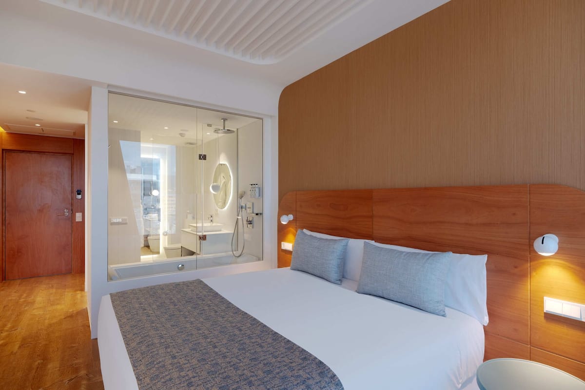 A modern hotel room with a large bed, a glass shower enclosure, wooden paneling, and a wooden door.