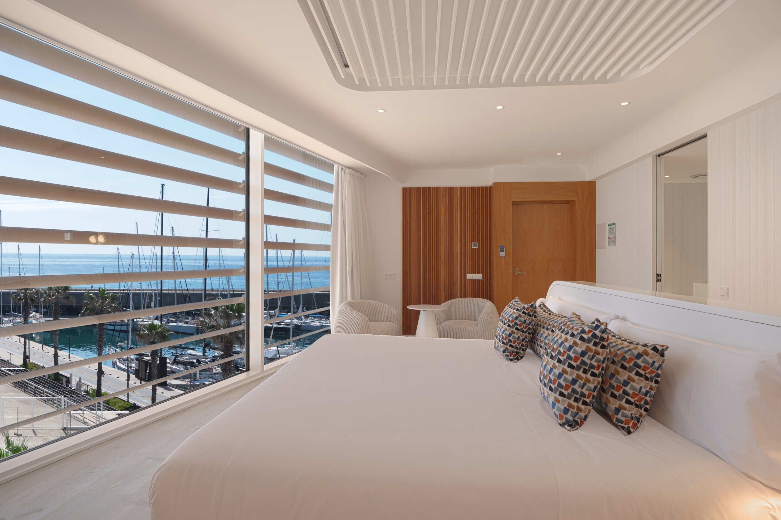 A modern bedroom with a large bed, a wooden wardrobe, a small table, and a view of the marina through floor-to-ceiling windows.