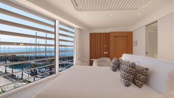 A modern bedroom with a large bed, a wooden wardrobe, a small table, and a view of the marina through floor-to-ceiling windows.