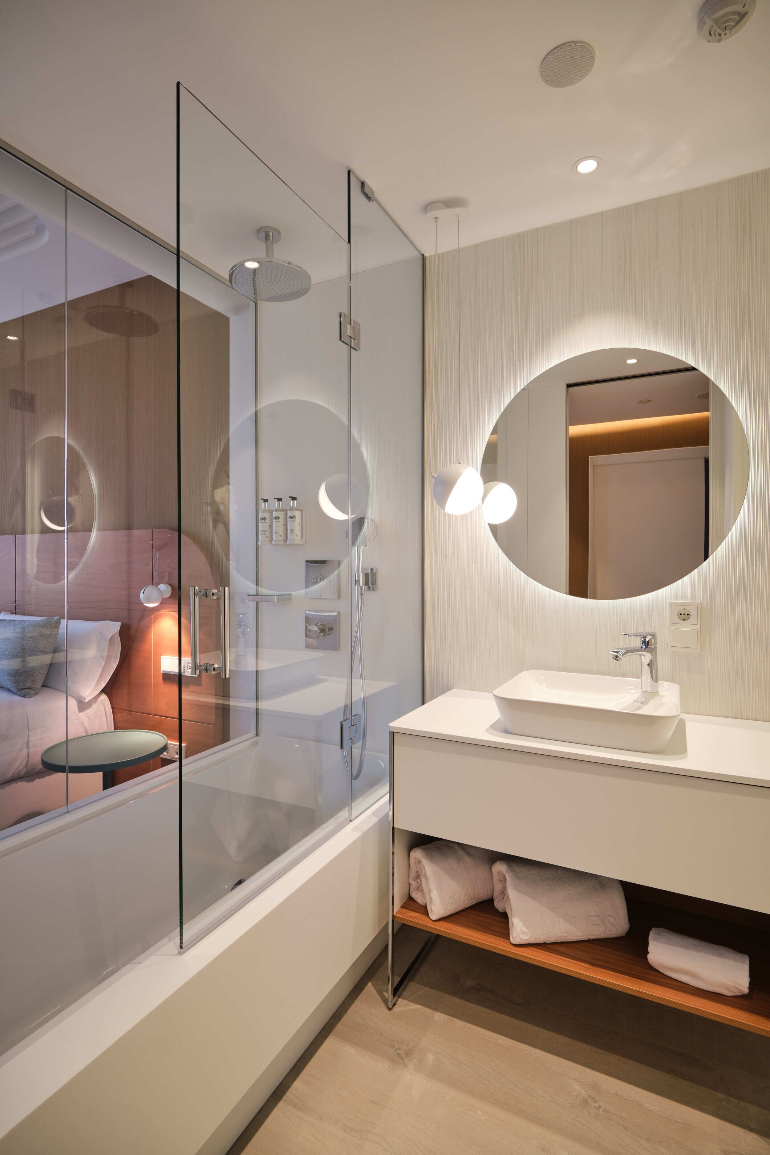 A modern bathroom with a glass shower enclosure, a white sink, and a round mirror with integrated lighting.