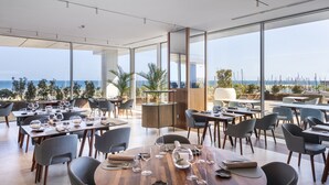 A modern dining area with tables set for a meal, large windows offering a view of the sea, and a wooden accent wall. - Hotel Marina Badalona (Badalona)