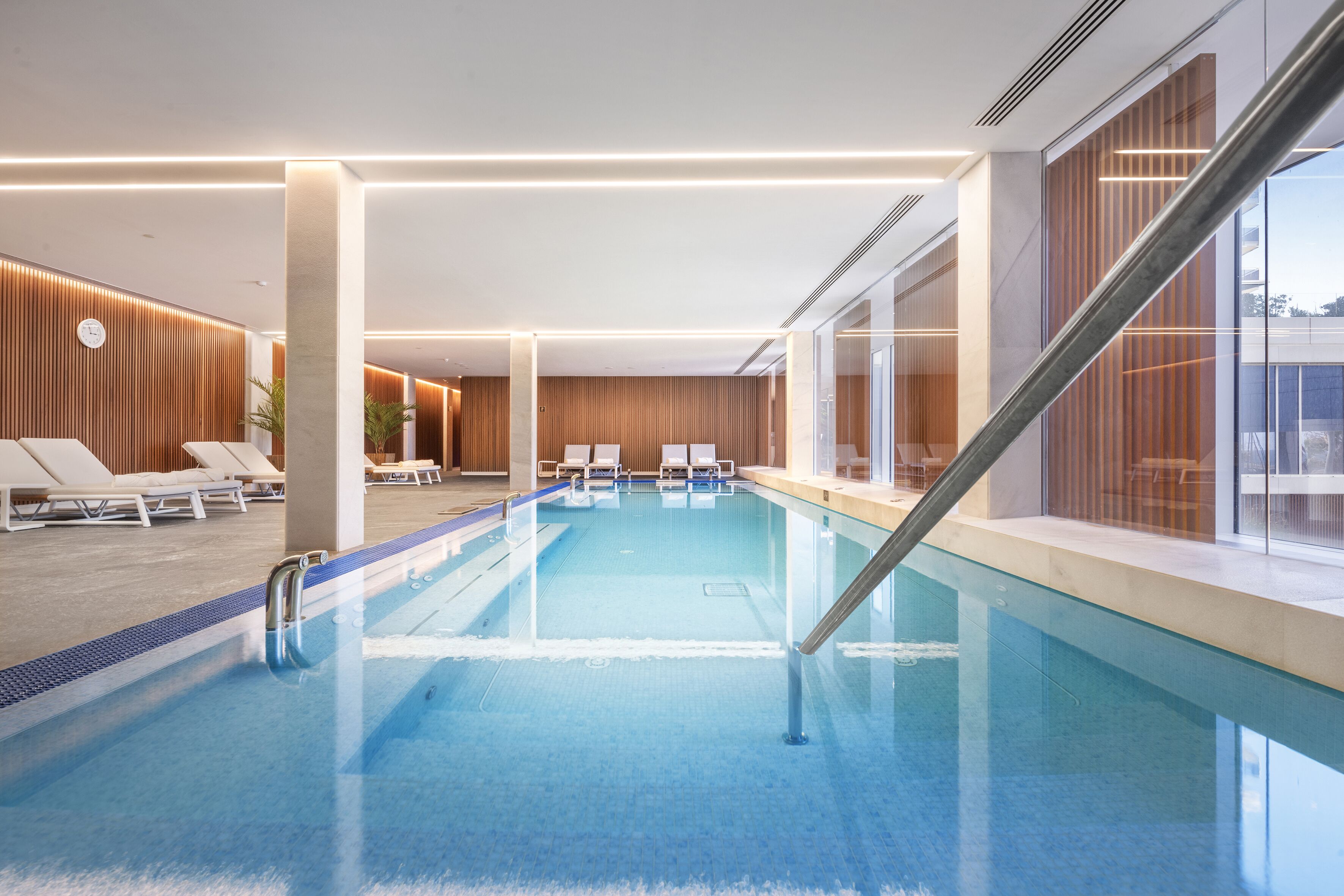 A modern indoor swimming pool with clear water, lounge chairs, and a wooden wall design.