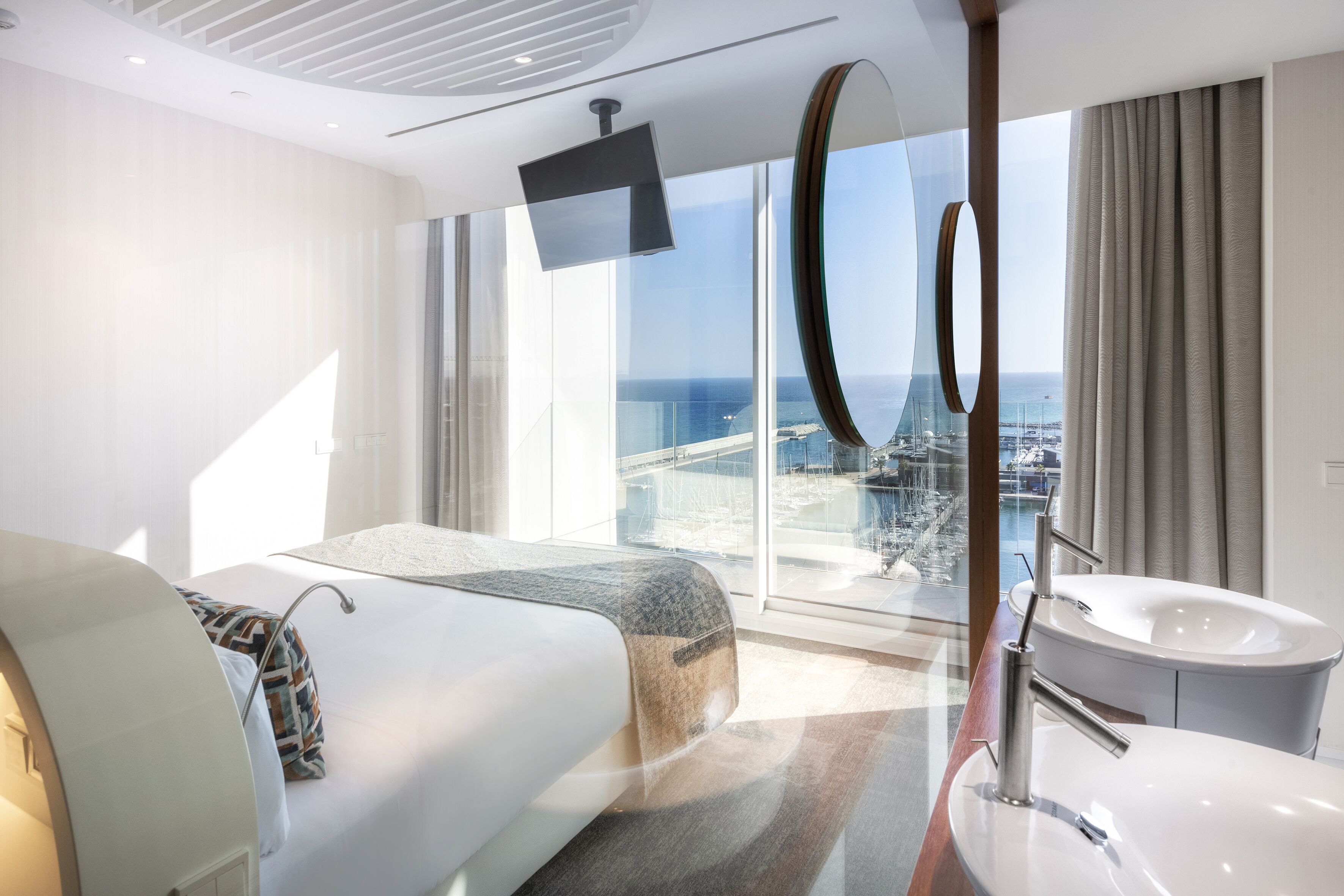 A modern hotel room with a large bed, a bathroom with a bathtub, a TV mounted on the wall, and a view of the city and sea through large windows.