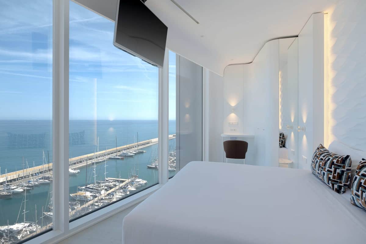 A modern hotel room with a large bed, a chair, and a view of a marina with boats.