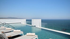 A rooftop infinity pool with clear blue water, white lounge chairs, and a glass railing overlooking the ocean. - Hotel Marina Badalona (Badalona)