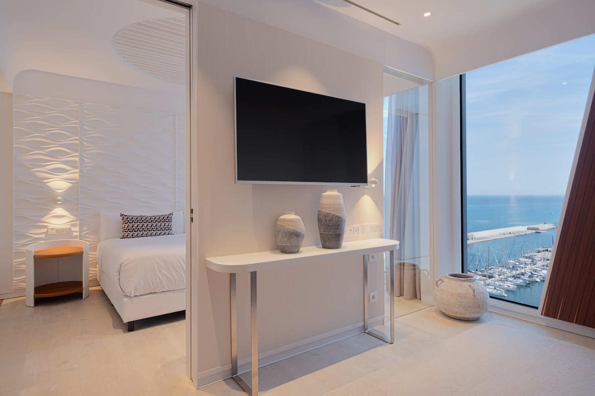 A modern bedroom with a large TV, a bedside table with decorative vases, a bed with patterned pillows, and a view of the sea through a large window.