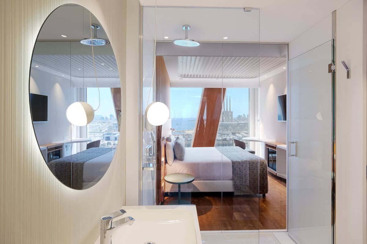 A modern hotel room with a large mirror, a bed with a city view, a small table, and a bathroom with a sink and shower.