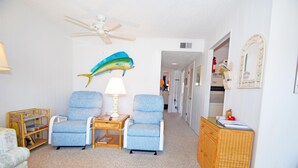 A living room with two blue armchairs, a small table with a lamp, a ceiling fan, a wall-mounted fish decoration, a wicker side table, and a wicker cabinet. - Bridgepoint II 5D-Bayfront 94th St, Free WiFi, Pool, W/D, AC (Ocean City)