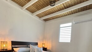 A bedroom with a bed, wooden ceiling beams, a window with blinds, and a nightstand with a lamp.