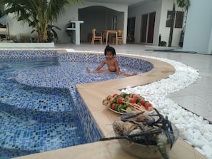 A child sitting in a circular pool with mosaic tiles, surrounded by white pebbles and a bowl of seafood. - Mango Groove Residency (Preah Sihanouk)