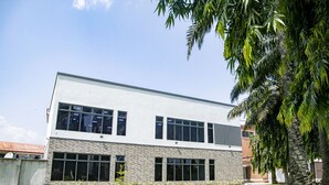 A modern two-story building with large windows and a flat roof, surrounded by a well-maintained lawn and palm trees.
