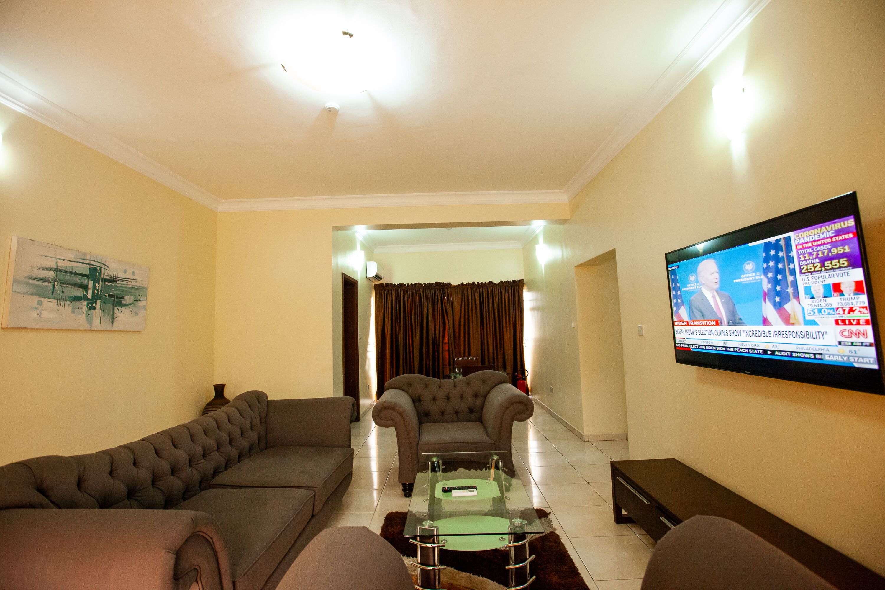 A living room with a sofa, armchair, and a glass coffee table. A flat-screen TV is mounted on the wall displaying a news channel.