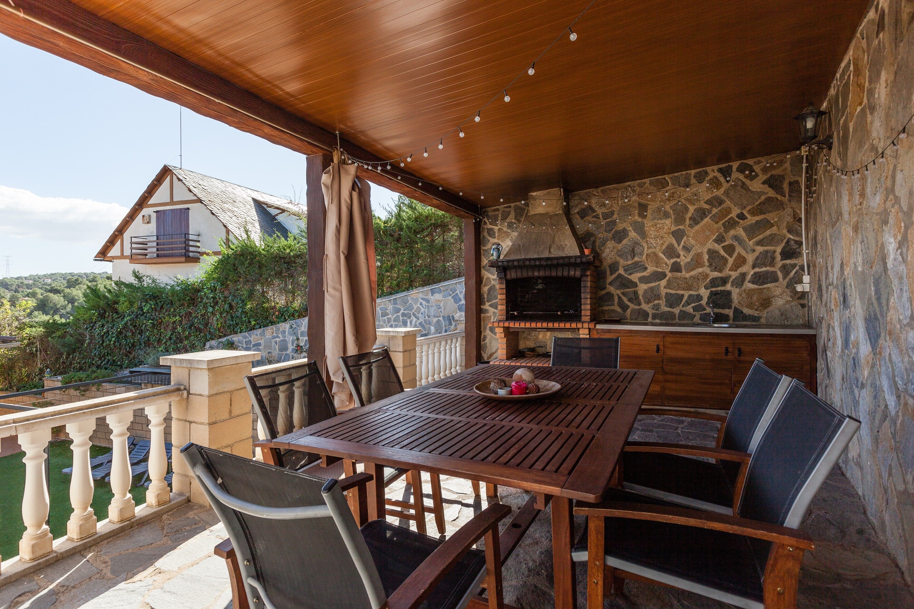 An outdoor patio with a wooden table and chairs, a stone fireplace, and a view of a house in the background.
