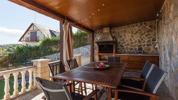 An outdoor patio with a wooden table and chairs, a stone fireplace, and a view of a house in the background.