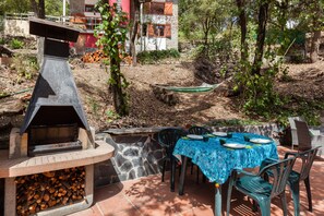 Ein Außenbereich mit Grillplatz, einem Steingrill, einem Tisch mit blauer Tischdecke und Stühlen.