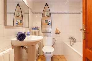 Ein Badezimmer mit weißem Waschbecken, WC und Badewanne. Zwei Spiegel mit Zierrahmen, eine Holztür und eine kleine Ablage mit Toilettenartikeln sind vorhanden.