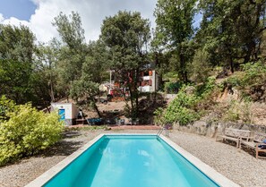 A swimming pool with a stone deck, surrounded by trees and a small building with a red roof.