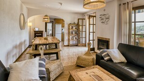 A living room with a wooden dining table, a black leather sofa, a woven pouf, and a hanging wicker light fixture. - Villa "Castell Mirto" with Private Pool, Terraces & Wi-Fi (Calonge)
