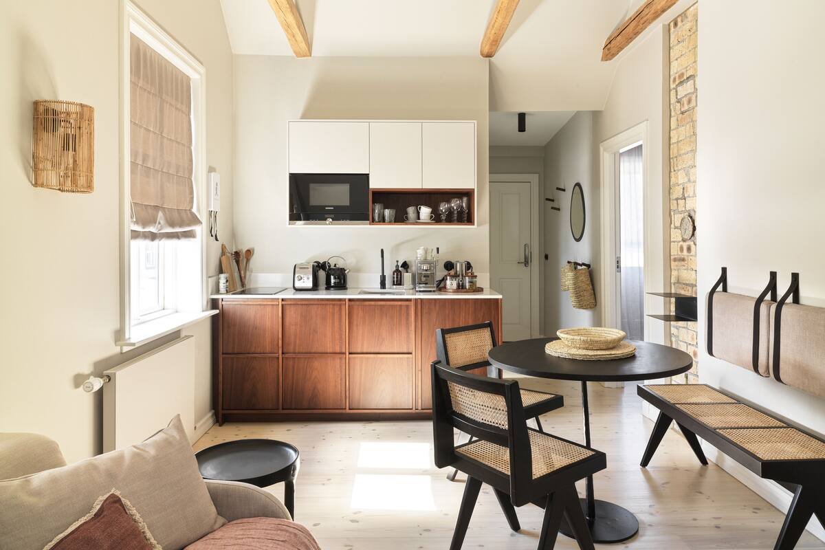 A modern kitchen with wooden cabinets, a microwave, and a coffee machine. There is a dining area with a round table and chairs.