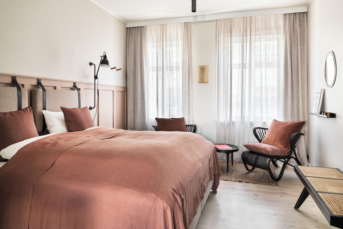 A hotel room with a large bed, a small table, a chair, and a window with sheer curtains.