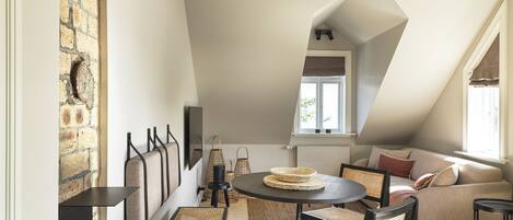 Une chambre à coucher confortable aménagée en mezzanine, avec un plafond en pente, un coin repas doté d’une table ronde et de chaises, et une grande fenêtre laissant entrer la lumière naturelle.