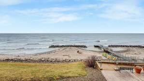 Une plage bordée d’une côte rocheuse, avec une jetée et une zone herbeuse où se trouvent un banc et des plantes en pot.