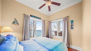 Une chambre à coucher avec un lit, un ventilateur de plafond, deux fenêtres avec des rideaux, une lampe et une vue sur l’extérieur.
