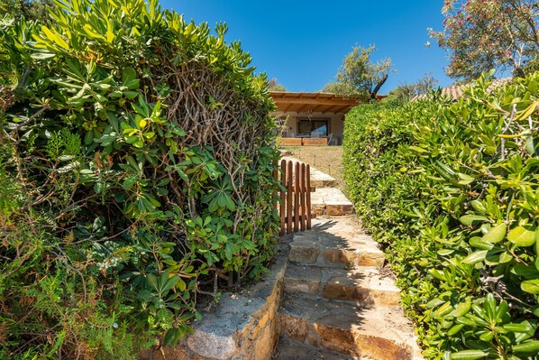 Un chemin de pierres mène à un bâtiment entouré d’une clôture en bois et bordé de chaque côté d’une végétation luxuriante.