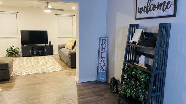 A modern living room with a flat-screen TV, a grey sofa, a wooden floor, and a 'welcome' sign.