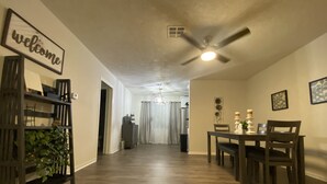 A spacious hallway with a wooden floor, a ceiling fan, and a dining area with a table and chairs. A 'welcome' sign is visible on the wall.