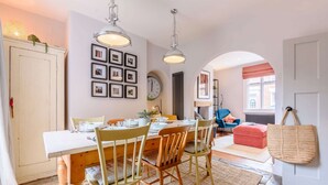 Un comedor con una mesa de madera, sillas e iluminadores colgantes. Hay un reloj en la pared y cuadros enmarcados sobre la mesa.