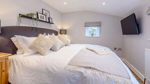 Un dormitorio con una cama grande, una mesita de noche, un televisor montado en la pared, una ventana con vistas y un estante con objetos decorativos.