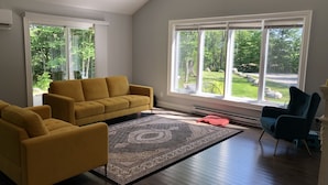 Un salón espacioso con un sofá amarillo, un tapete estampado y vistas a un paisaje verde a través de amplias ventanas.