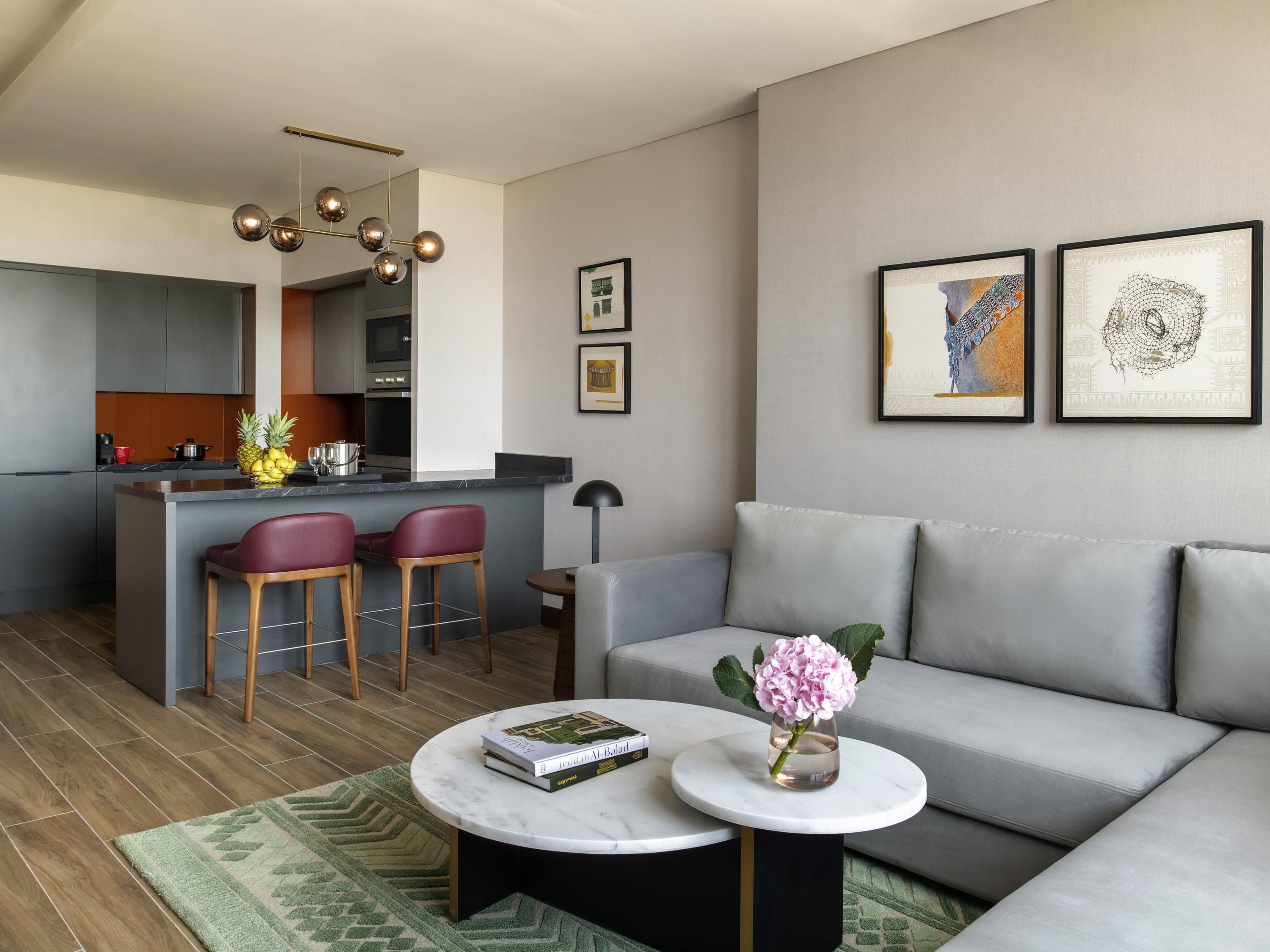A modern living room with a grey sofa, a round coffee table with a book and a vase of flowers, a kitchen area with a bar, and two framed artworks on the wall.