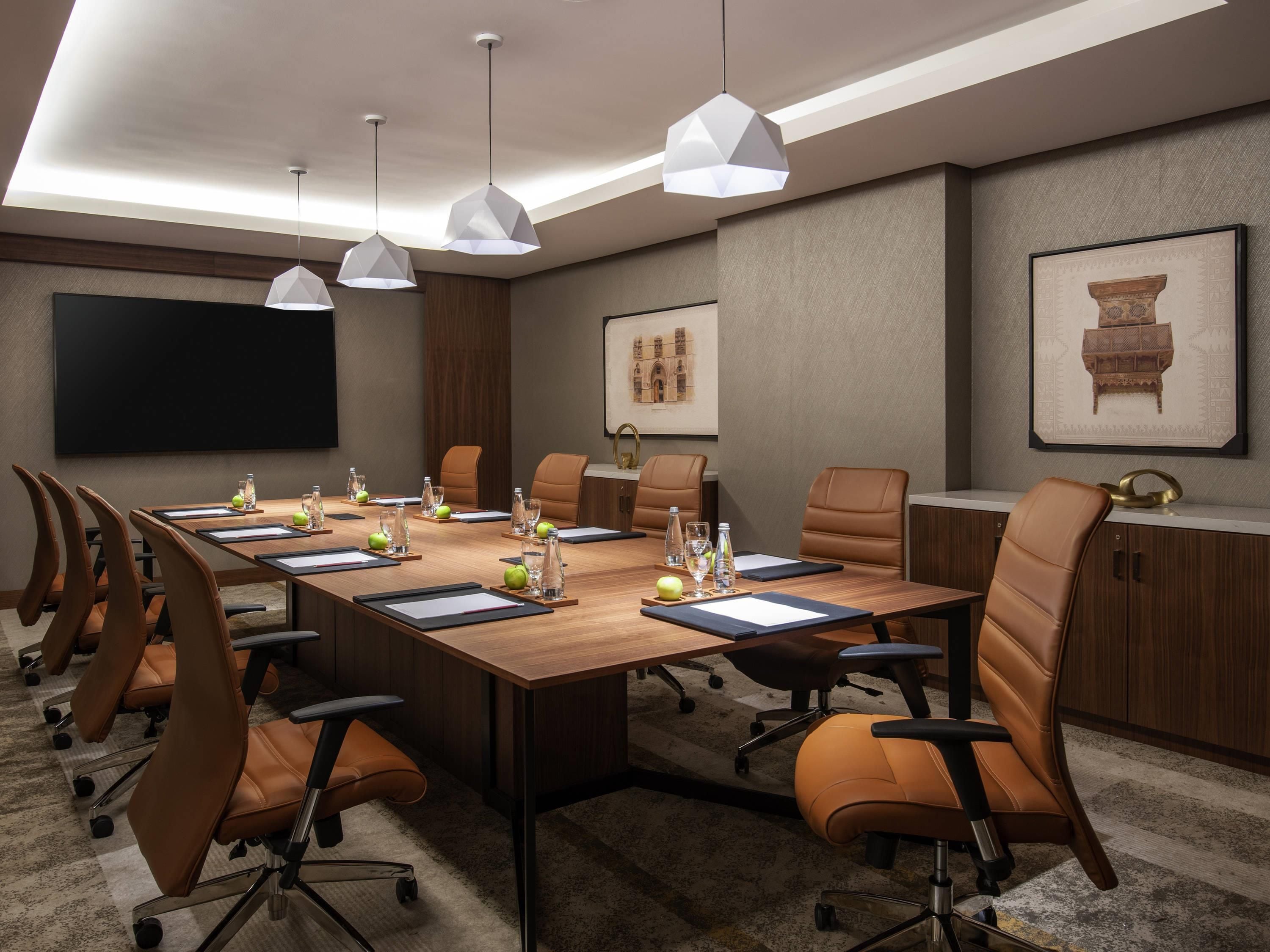 A conference room with a long table, brown leather chairs, white pendant lights, and a large flat-screen TV.