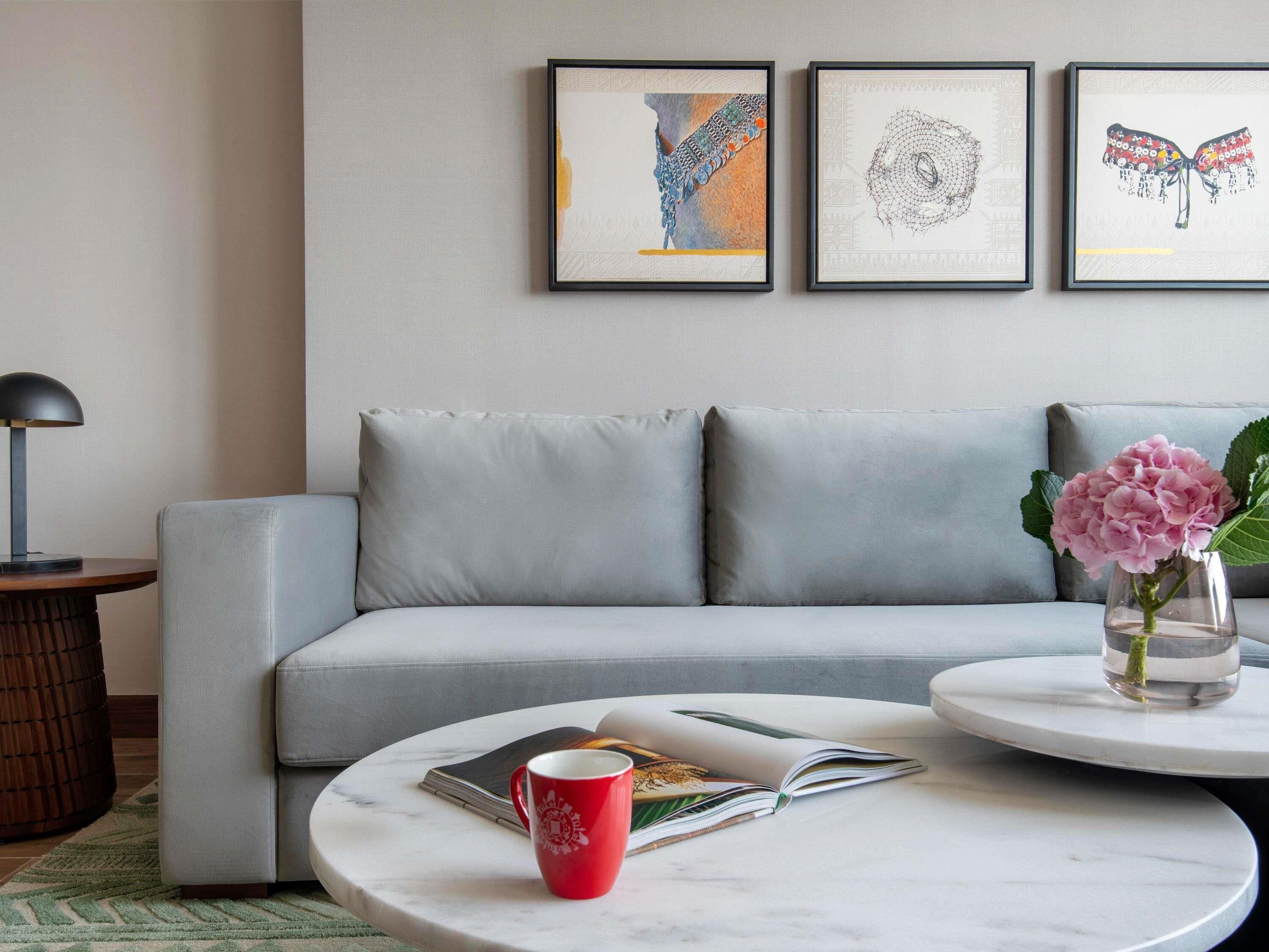 Una sala de estar moderna con un sofá gris, una mesa redonda de centro con un libro y una taza roja, un jarrón con flores rosas, y tres cuadros en la pared.