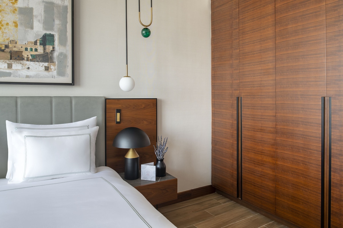 A neatly made bed with white linens and a gray headboard, a bedside table with a lamp and a vase, and a wooden wardrobe with a mirror.