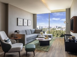 Ein modernes Wohnzimmer mit einem grauen Sofa, einem grünen Hocker, einem runden Couchtisch und Blick auf die Stadt durch große Fenster.